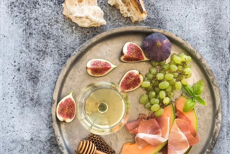 Wine appetizer set. Glass of white wine, honeycomb with drizzlier, figs, grapes, melon and prosciutto on silver tray over rustic grunge surface. Top view