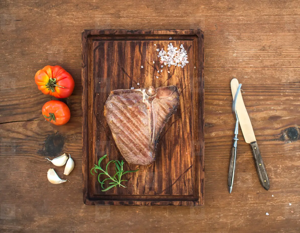 Cooked meat t bone steak on serving board with garlic cloves  tomatoes  rosemary and spices over rustic wooden background