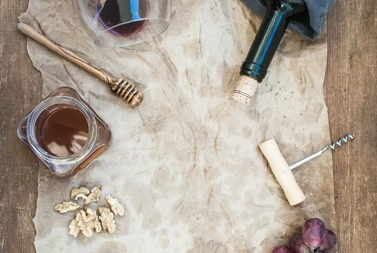 Wine and appetizer set with copy space in center. Glass of red wine, bottle, corkscrewer, blue cheese, grapes, honey, walnuts on oily craft paper over rustic wooden table, top view.
