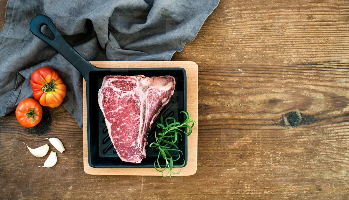 Raw uncooked meat t bone steak with garlic  heirloom tomatoes and rosemary in cooking pan over rustic wooden background
