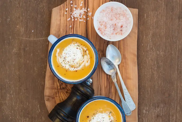 Homemade pumpkin cream soup in enamel mugs with herbs and fresh bread slices on olive serving board over rustic wooden background