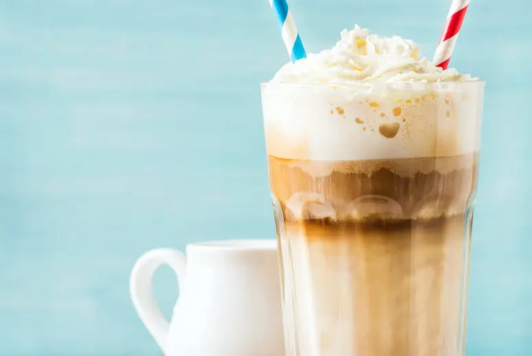 Latte macchiato with whipped cream in tall glass , two straws and pitcher on wooden board over blue painted wall background, copy space