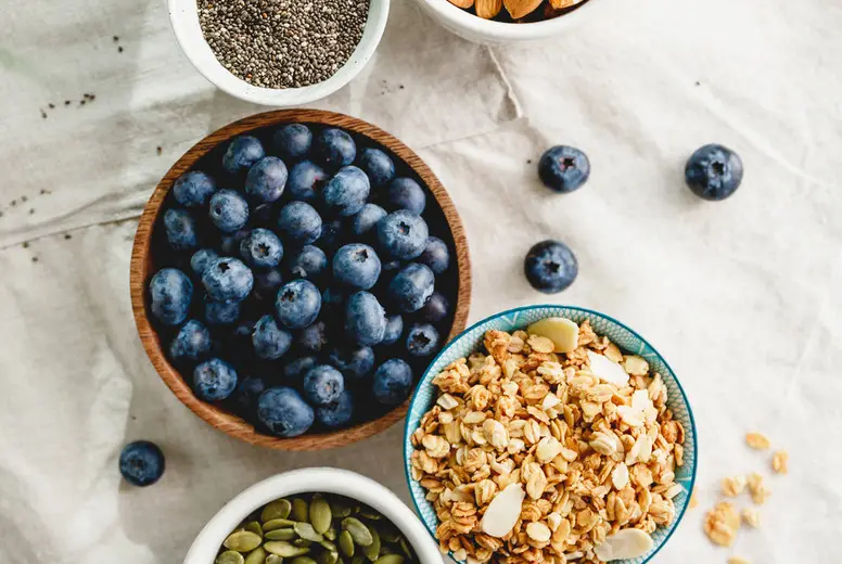 Top view of breakfast ingredients like cereal, almond, blueberry, pepitas and chia seeds on a linen cloth decorated with palm leaf.