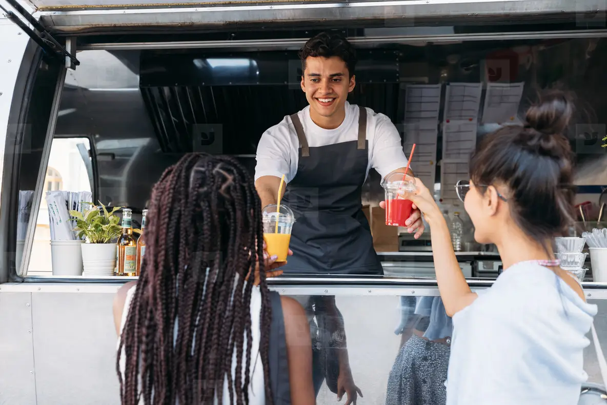 Back view of two females buying drinks from a male owner at a food truck  Smiling entrepreneur giving drinks to clients