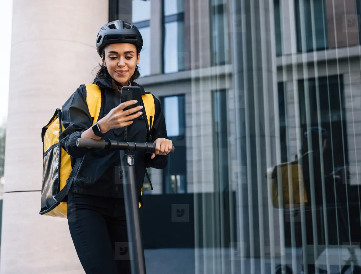 Smiling delivery woman with backpack looking on smartphone leaning on a handlebar of electric push scooter