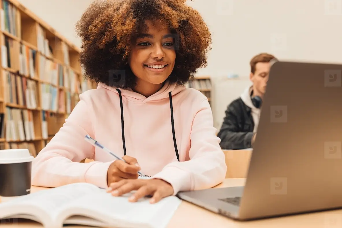 Smiling girl writing in a book while sitting at desk with laptop  Female student studying at college library