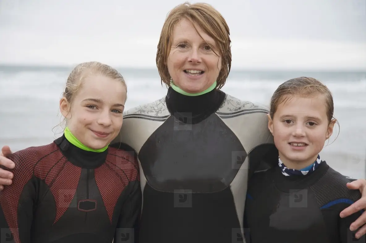 Woman and two girls wearing surfing wetsuits standing on a beach smiling