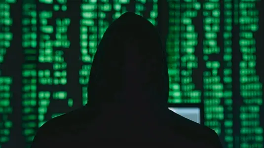 hooded figure stands before glowing green code  symbolizing cyber threats
