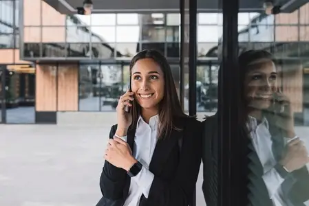 Smiling female in black formal wear leaning business building and talking on mobile phone  Female office worker talking on mobile phone during a break