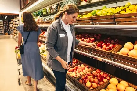 Female grocery store worker stands by the shelves as buyer with a cart walks past