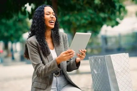 A Smiling Woman Delightfully Enjoys Using a Tablet While Relaxing in an Urban Park Setting