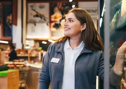 Young smiling female grocery store worker standing at the fridge and looking away