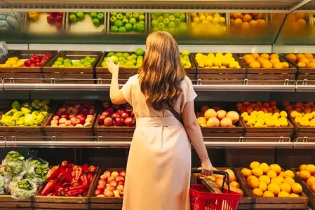 Rear view of a woman in casual clothes standing at shelves with fruits and vegetables holding shopping basket