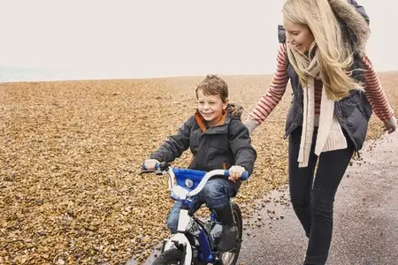 Happy mother helping son learn how to ride bicycle on path along rocky beach