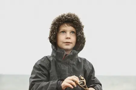 Curious boy in fur lined jacket holding slingshot on winter beach