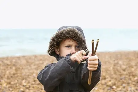 Cute boy in fur lined jacket playing with slingshot on rocky beach