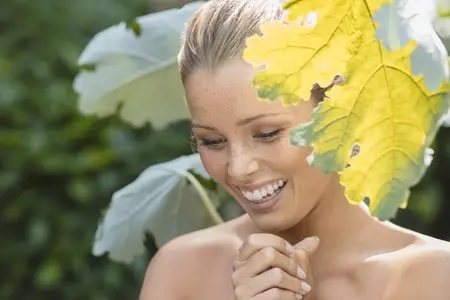 Beautiful young woman with bare shoulders smiling and looking down among green leaves in garden