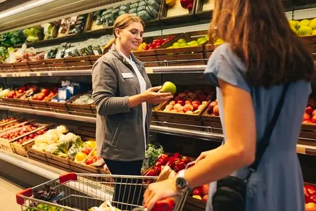 Smiling female worker standing at shelves with fruits and vegetables and showing an apple to a customer