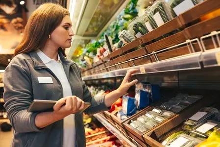 Female grocery store worker checking prices on shelves in a grocery store and holding a digital tablet
