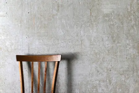 A minimalistic photo of a wooden chair with vertical slats placed against a textured concrete wall
