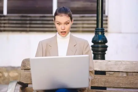 A surprised woman in casual clothes sits on a park bench using her laptop outdoors