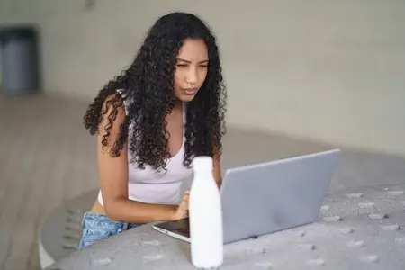 A focused  young woman is diligently working on her laptop with a bottle visible in the background