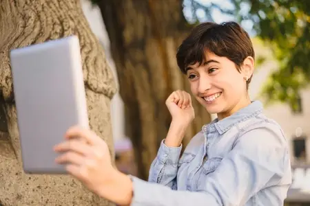 A Joyful Young Woman Happily Taking a Selfie Outdoors with Her Tablet in Bright Daylight