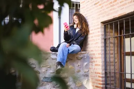 A casually dressed young woman is sitting on a wall in a relaxed posture  using her smartphone