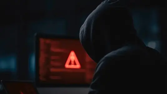 Hacker in dark room with hooded sweatshirt  red warning symbol on computer screen  cybercrime