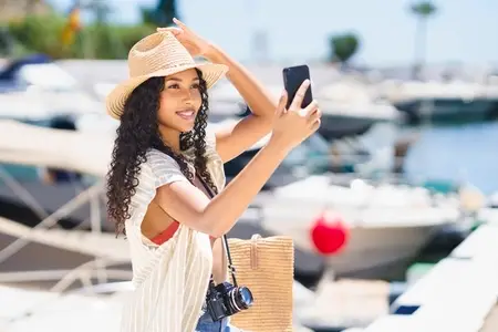 A cheerful young woman takes a selfie at a vibrant marina on a bright sunny day