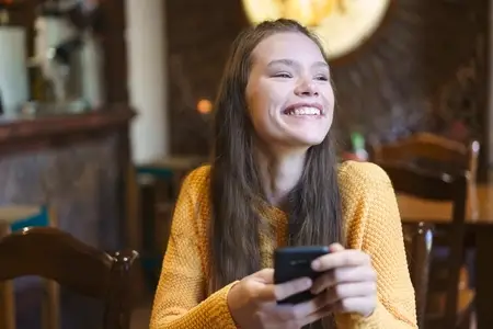 A cheerful young woman is smiling widely while she is using her smartphone in a cozy cafe