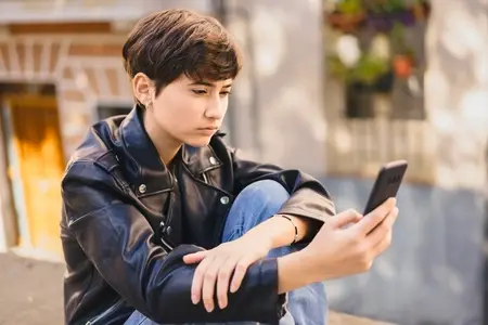 A young teenager with a smartphone is seated in an urban setting  surrounded by a lively cityscape
