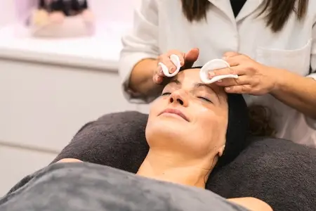 Crop beautician removing facial product from face with cotton pads