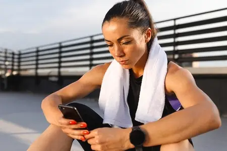 Slim athlete with a white towel on her neck using a phone while taking a break during a workout