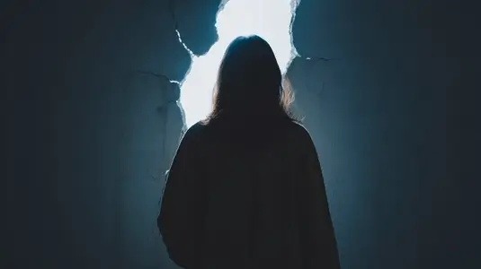 Silhouette person standing in dark room facing cracked wall with light shining through opening
