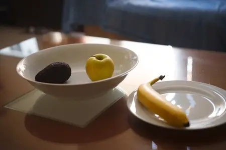 Bowl with apple and avocado beside banana plate atop glossy table  soft sunlight spilling over ceramic  cozy living room blur  fresh organic snack for calm morning