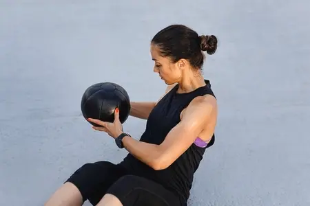 Muscular young female with medicine ball practicing outdoors  Woman in black fitness attire exercising using medicine ball while sitting