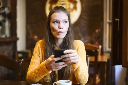 A Thoughtful and Young Woman Happily Enjoying a Delicious Coffee While Using Her Smartphone